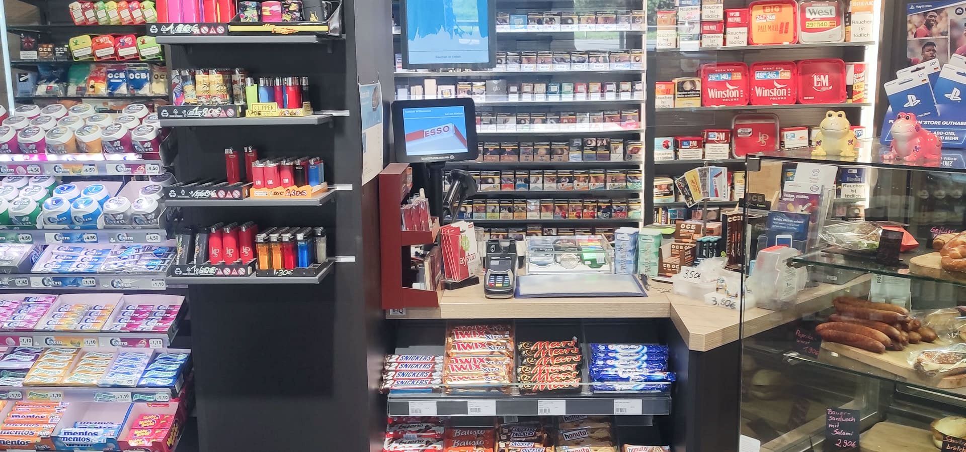 Verkaufsraum eines Kiosks mit Regalen voller Süßigkeiten, Tabakwaren und einem Tresen mit Backwaren und einer Kasse